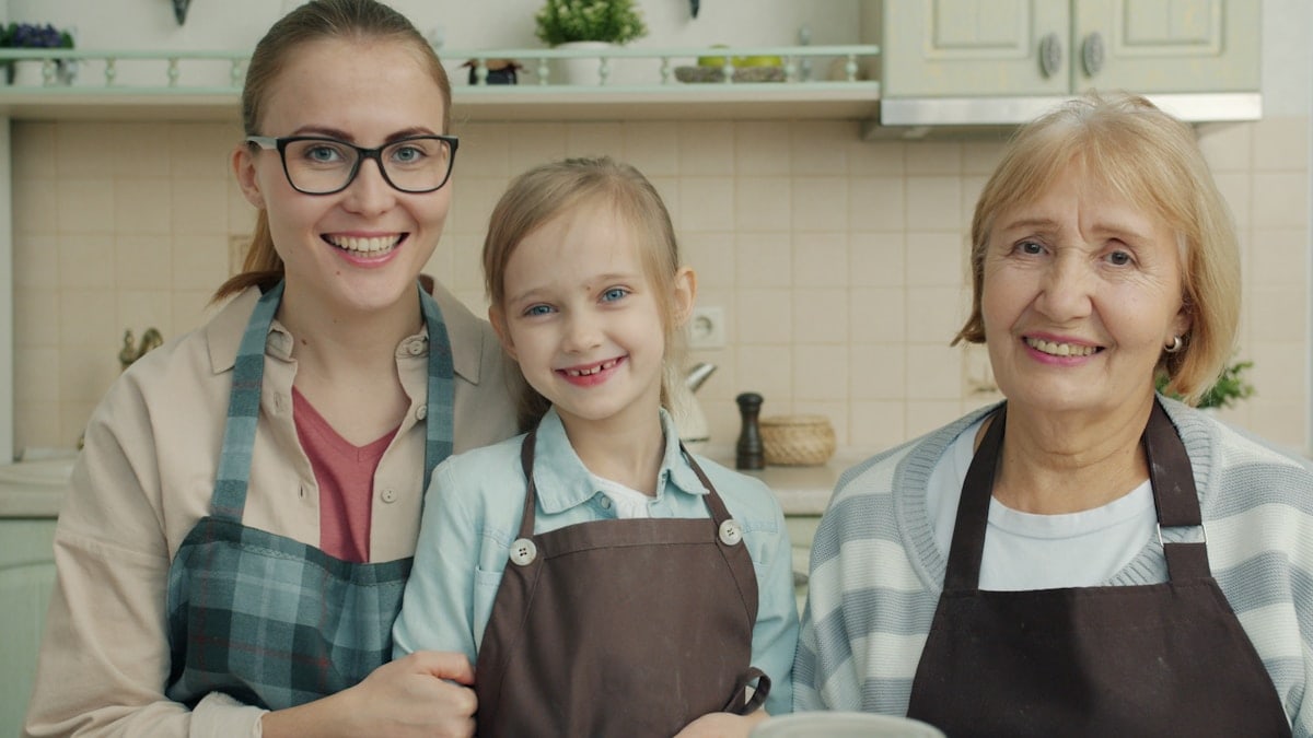 Portrait of happy family women and small girl in aprons smiling sitting in kitchen together looking at camera. Lifestyle, cookery and people concept.
